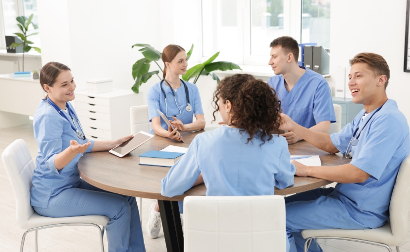 Năm nhân viên y tế mặc bộ đồng phục (scrubs) màu xanh dương ngồi quanh bàn tròn gỗ, đang trao đổi. Một người cầm máy tính bảng, một người cầm bút. Phòng làm việc hiện đại, ánh sáng tự nhiên từ cửa sổ
