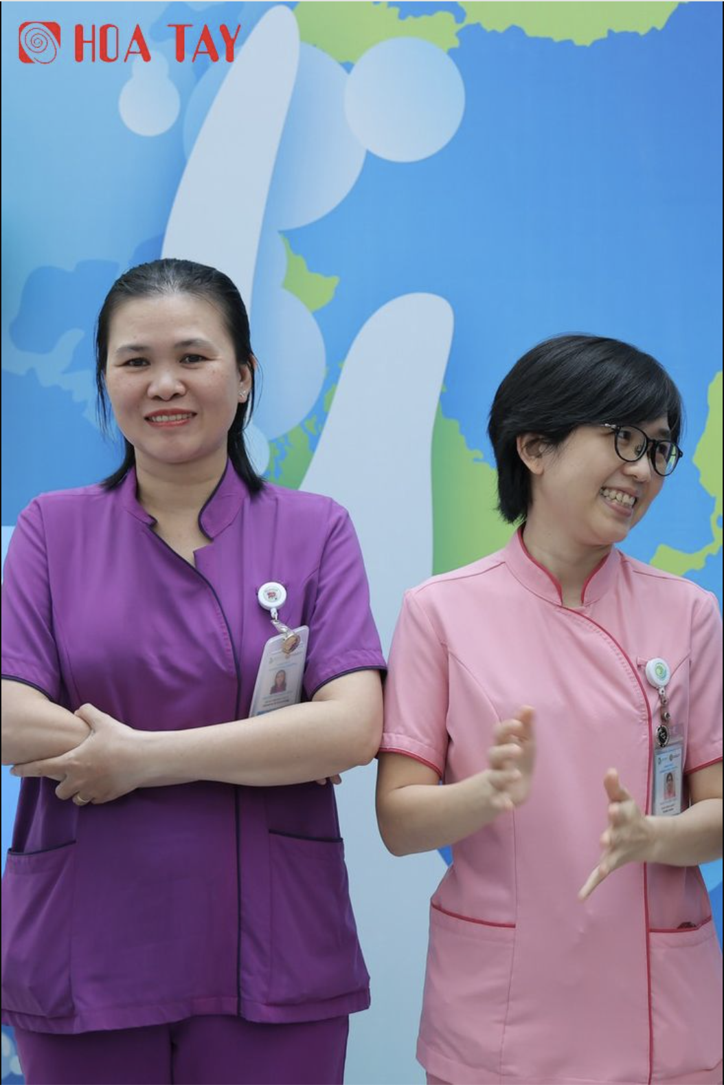Hai nữ nhân viên y tế (hai người) đang đứng, mặc áo đồng phục cổ tàu (mandarin collar) màu tím và hồng, tay áo ngắn, có túi hông, đeo thẻ tên trước ngực, phông nền xanh dương có họa tiết bản đồ, ánh s