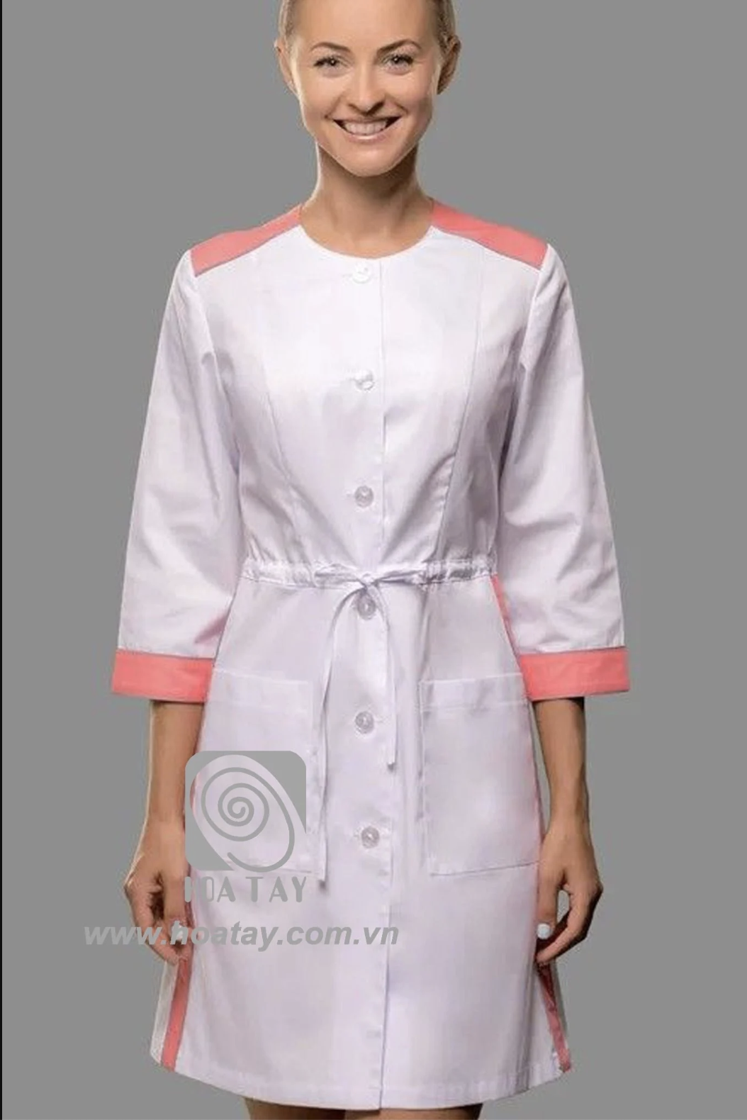 Một nữ nhân viên y tế đang đứng thẳng, mặc áo blouse trắng phối hồng (viền vai và tay áo), cổ tròn, tay lỡ, có dây rút eo (thắt lưng điều chỉnh) và hai túi trước, nền xám, ánh sáng studio.