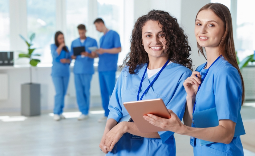 Bốn nhân viên y tế mặc bộ đồng phục (scrubs) màu xanh dương, hai người ở tiền cảnh cầm máy tính bảng và kẹp tài liệu, ba người ở hậu cảnh đứng trao đổi. Thiết kế cổ tròn, tay ngắn, chất liệu vải mềm,