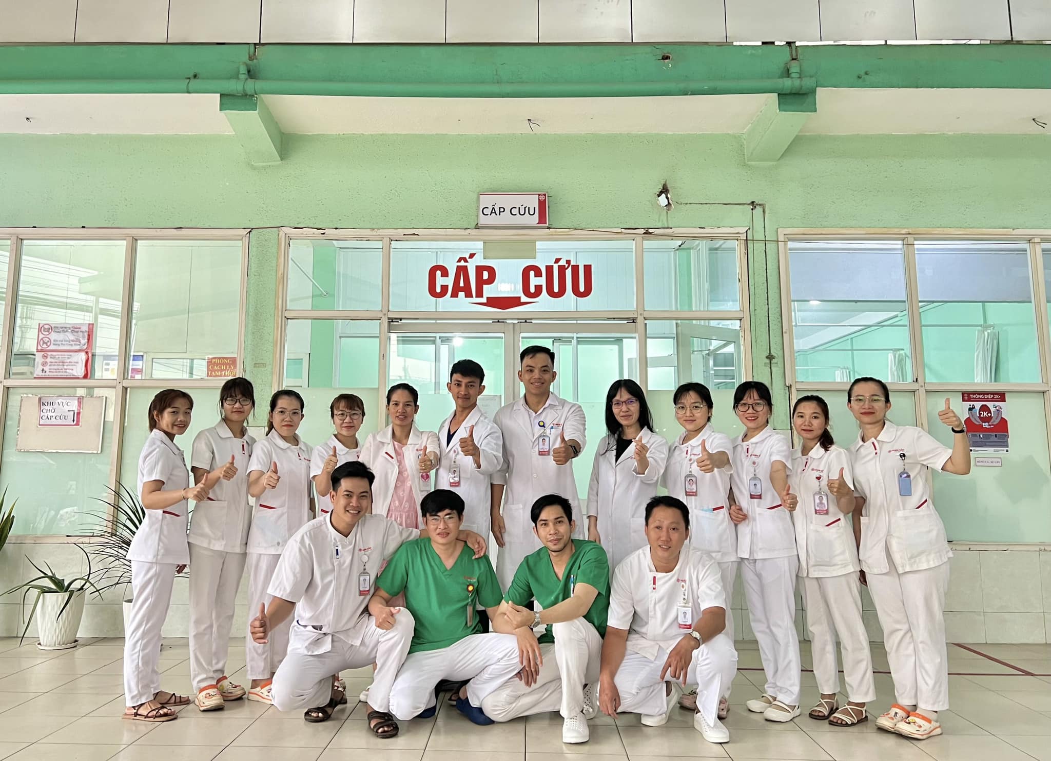 Nhóm 15 nhân viên y tế đang đứng và ngồi tạo dáng giơ ngón cái; mặc áo blouse trắng (áo khoác bác sĩ) hoặc áo scrub xanh (đồng phục phẫu thuật) vải cotton, cổ chữ V hoặc cổ bẻ, tay ngắn; bối cảnh hành