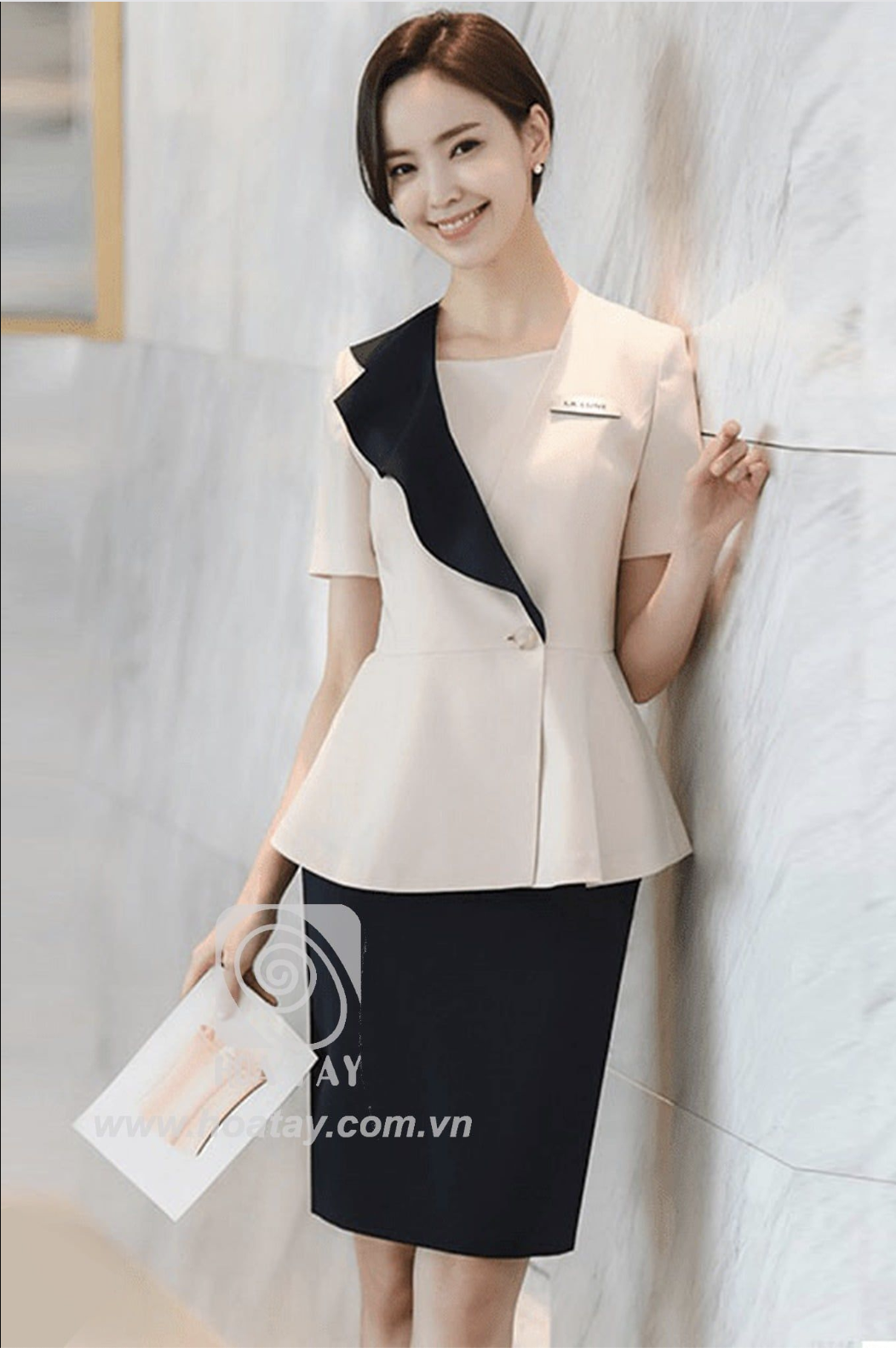 Một nữ nhân viên đứng tựa tường; mặc áo peplum (dáng xòe eo) màu kem phối ve áo đen, tay ngắn, cài một khuy, chất liệu vải đứng form; bối cảnh hành lang sáng sủa.