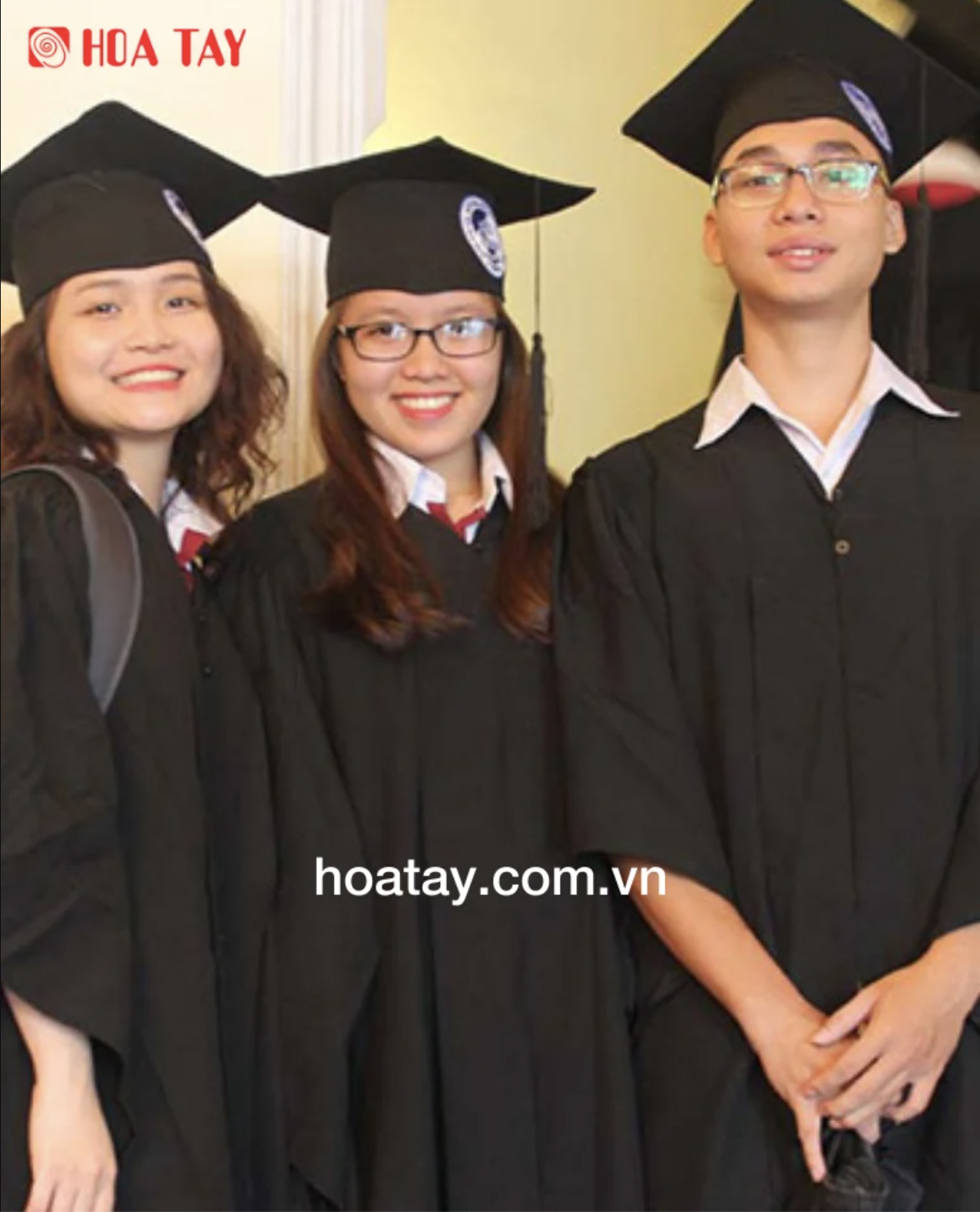 Ba sinh viên (ba người) đang mỉm cười, mặc lễ phục tốt nghiệp (áo cử nhân) màu đen, chất liệu vải trơn, mũ vuông có tua rua, cổ áo sơ mi trắng bên trong, bối cảnh trong nhà với ánh sáng vàng dịu.