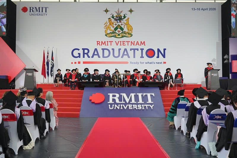 Hơn 20 sinh viên và giảng viên đang ngồi trên sân khấu lễ tốt nghiệp, mặc áo choàng (áo cử nhân) đen, mũ vuông (mũ tốt nghiệp) có tua rua, thảm đỏ dẫn lối, phông nền logo RMIT và huy hiệu trường, ánh