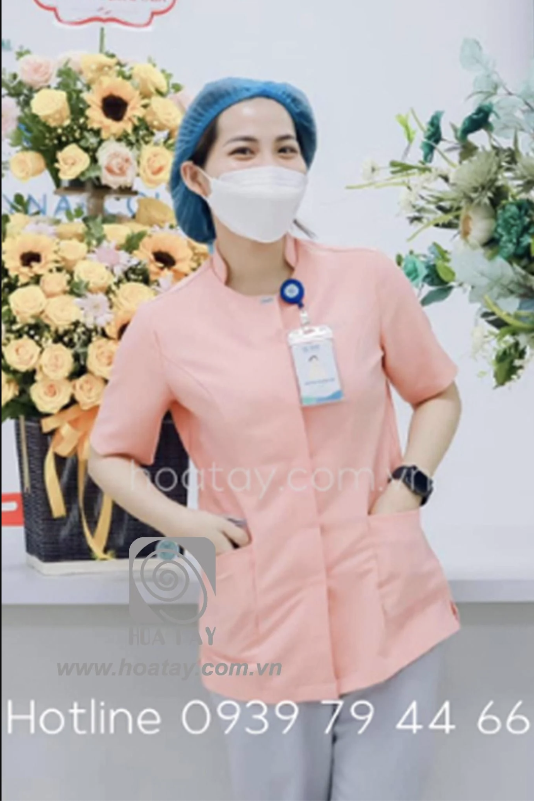 Một nữ nhân viên y tế đang đứng, mặc áo blouse cổ tàu (cổ đứng) màu hồng phấn, tay ngắn, hai túi hông lớn, chất vải phẳng phiu, đeo thẻ tên và mũ bao tóc xanh, bối cảnh phòng khám có lẵng hoa tươi, án