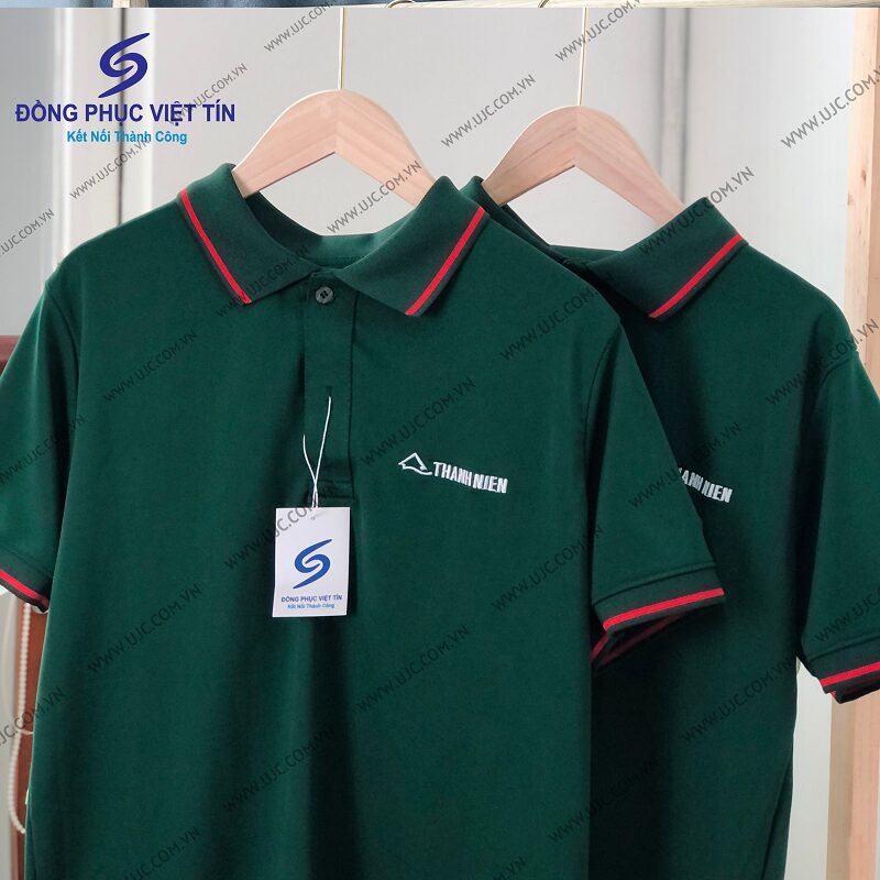 Hai chiếc áo polo màu xanh lá cây đậm với viền cổ và tay áo màu đỏ, có thêu logo 'THANH NIEN' trên ngực, treo trên móc gỗ trong không gian sáng sủa.