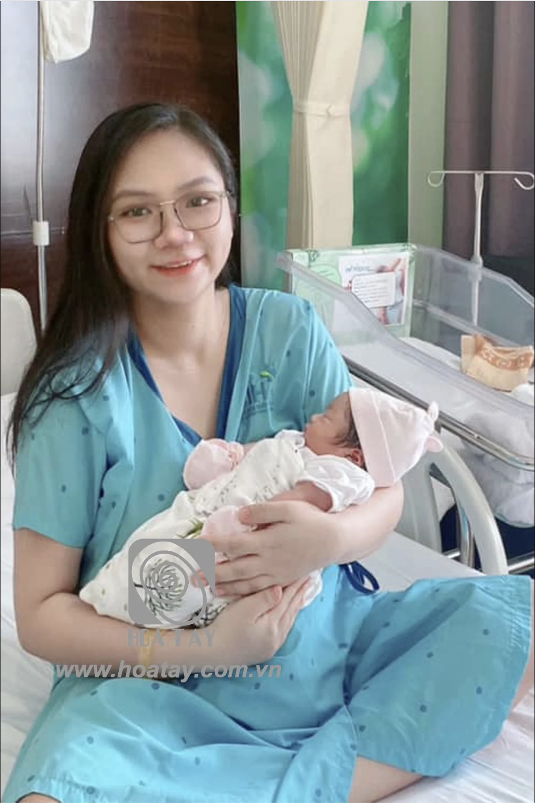 Một người mẹ và một trẻ sơ sinh (hai người) -> đang bế ẵm -> mặc bộ đồ bệnh nhân (scrubs) màu xanh lơ, chất vải cotton mềm, cổ chữ V, em bé quấn tã trắng, đội mũ hồng -> trong phòng bệnh viện, ánh sán