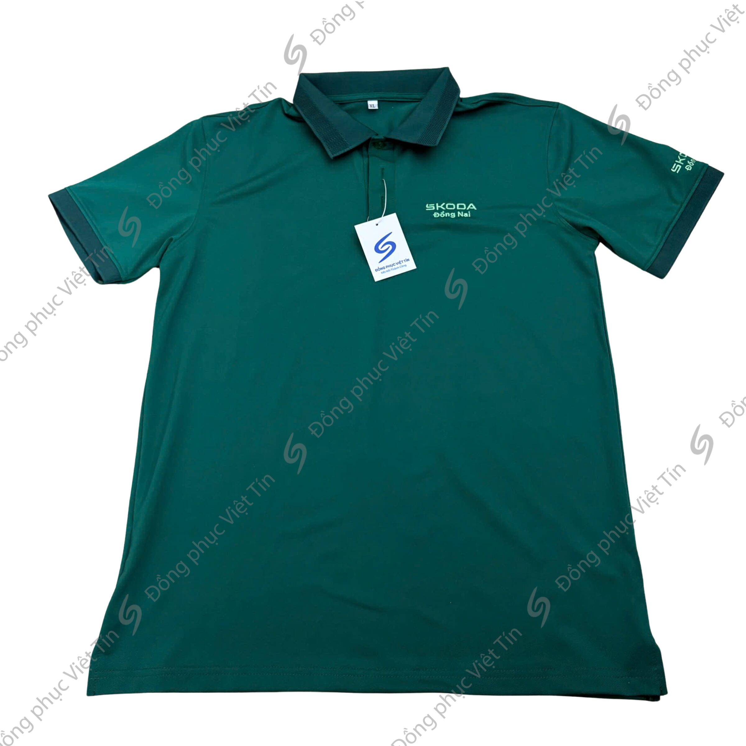 Áo polo màu xanh đậm, cổ bẻ, tay ngắn, có logo Skoda Đồng Nai thêu trước ngực và trên tay áo, kèm thẻ bài sản phẩm, đặt trên nền trắng phẳng.