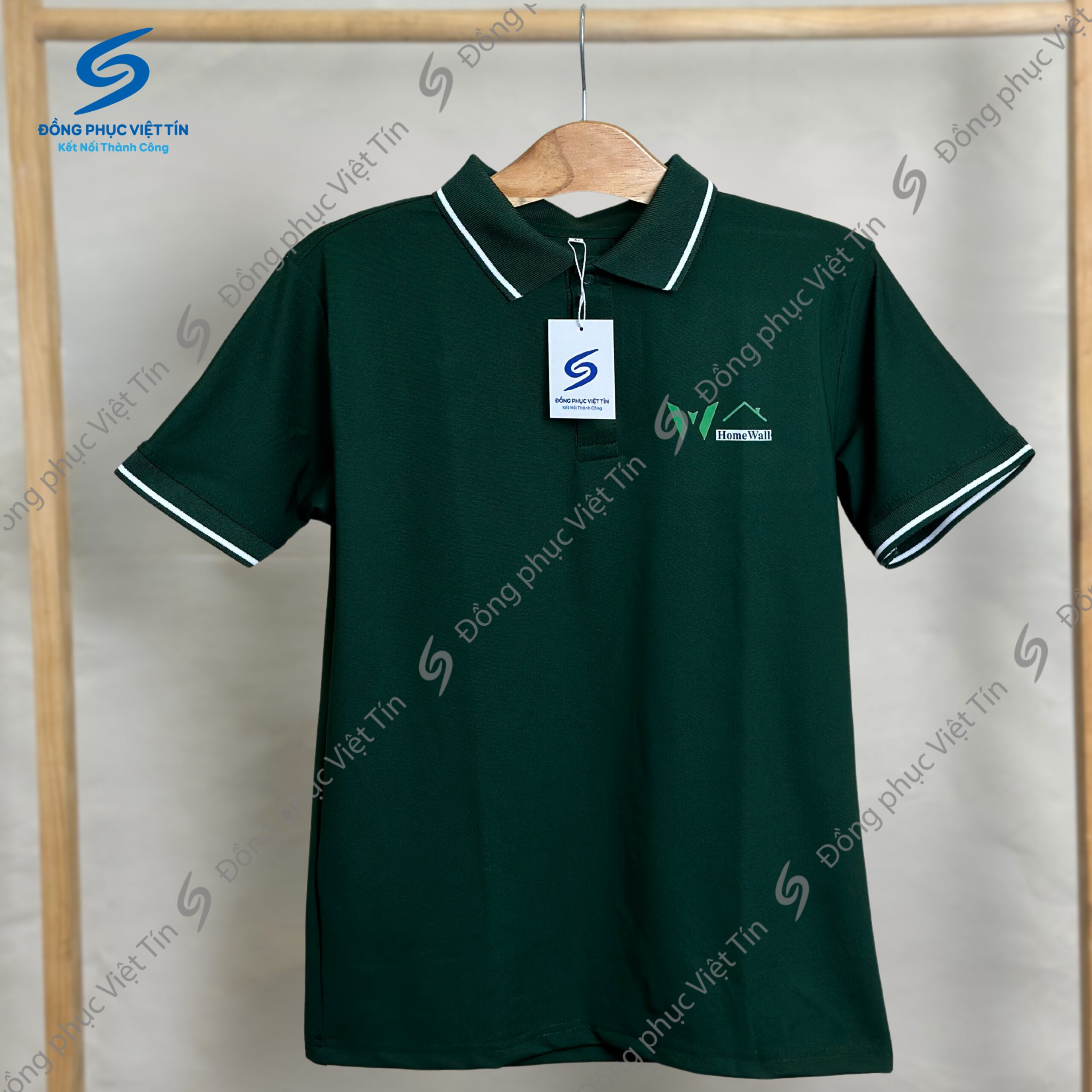 Áo polo màu xanh lá đậm, cổ và tay áo có viền trắng, treo trên móc gỗ. Áo có logo HomeWall thêu trước ngực và nhãn hiệu Đồng phục Việt Tín.