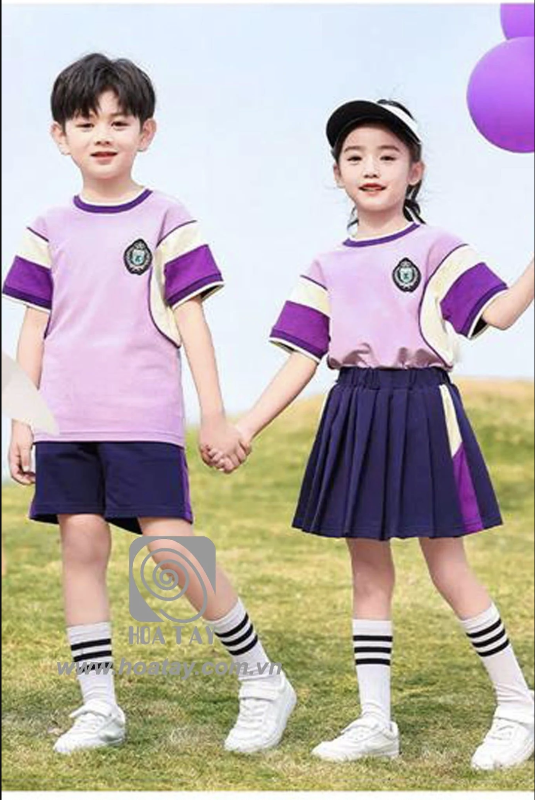 Hai học sinh (một nam, một nữ) đang nắm tay nhau; mặc áo thun cổ tròn phối màu tím-kem, quần short/chân váy xếp ly màu tím than, tất cao cổ; bối cảnh ngoài trời, ánh sáng tự nhiên.