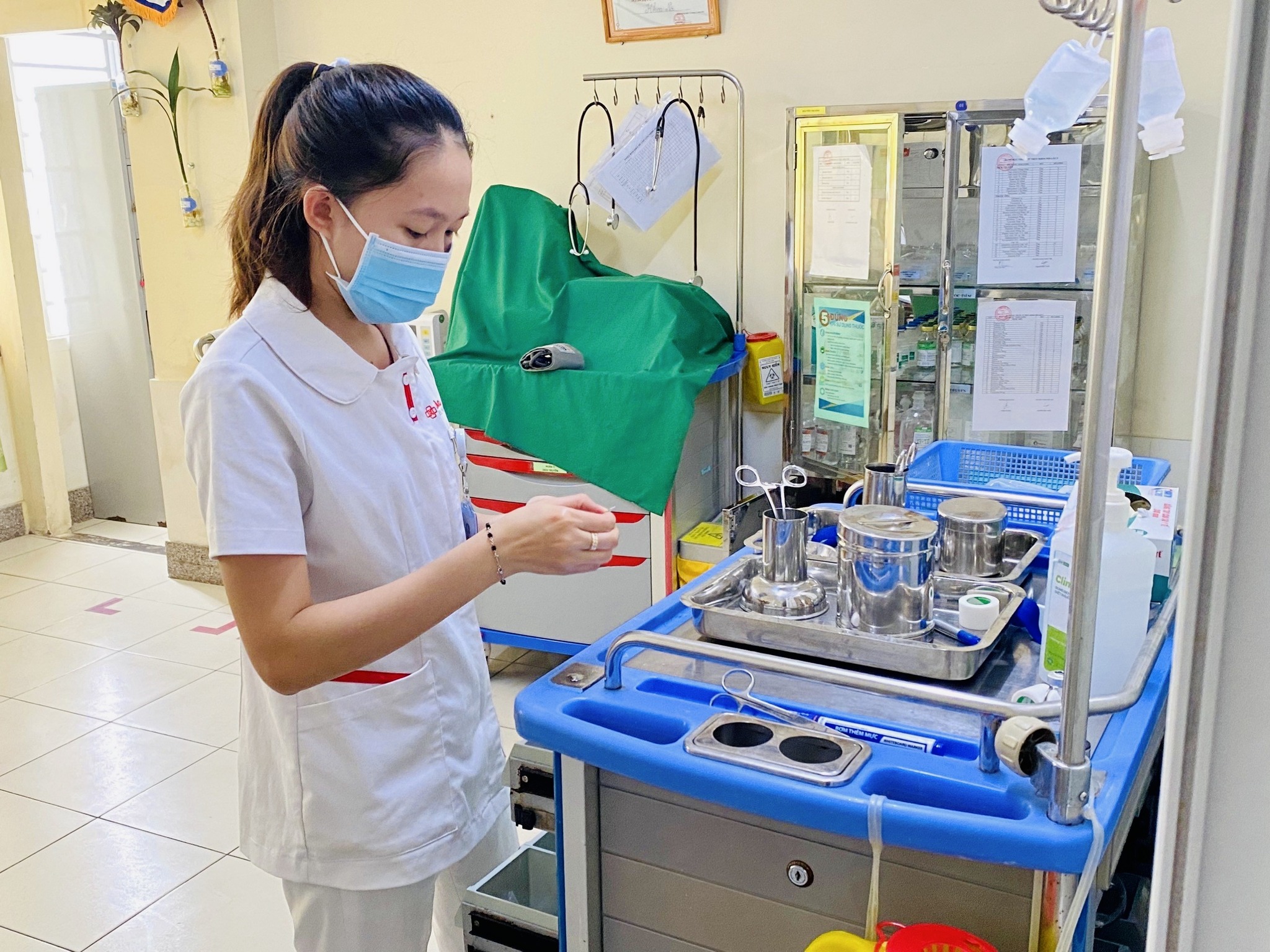 Một nữ điều dưỡng đang chuẩn bị dụng cụ y tế; mặc áo blouse trắng cổ bẻ (cổ áo sơ mi), vải cotton, viền đỏ ở túi, đeo khẩu trang y tế xanh, trong phòng bệnh viện ánh sáng tự nhiên.