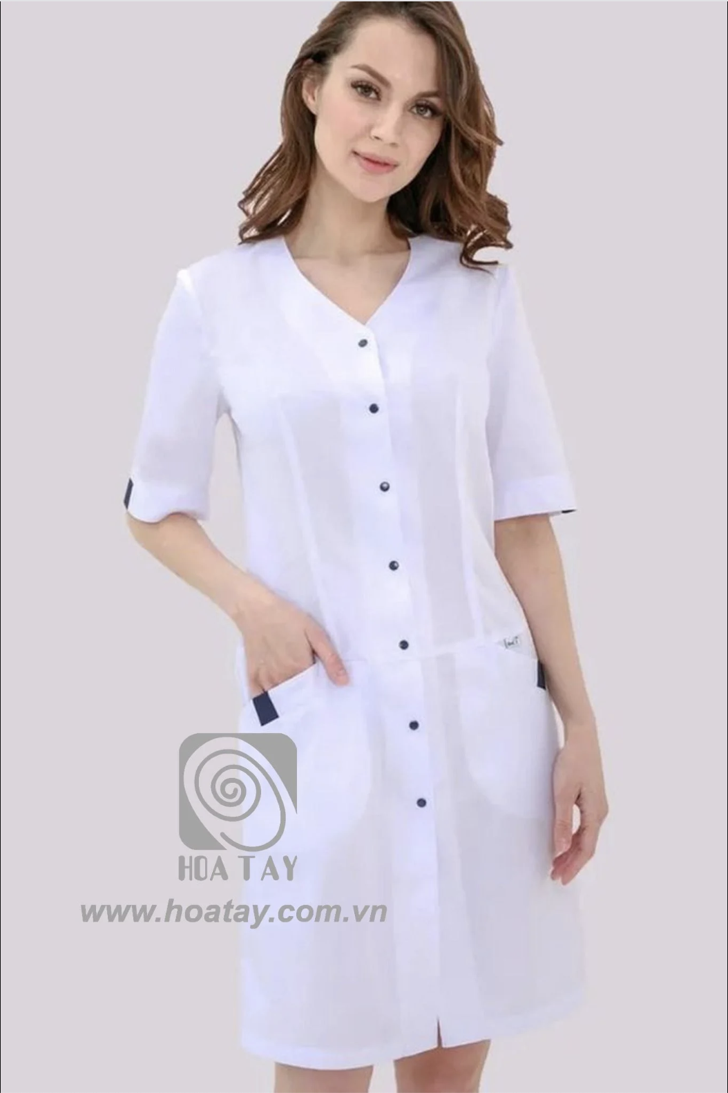 Một nữ nhân viên y tế đang đứng, mặc áo blouse trắng (áo khoác phòng thí nghiệm) tay lỡ, cổ chữ V, cài khuy dọc thân, có hai túi hông phối viền xanh đen, chất liệu vải phẳng mịn, phông nền studio sáng