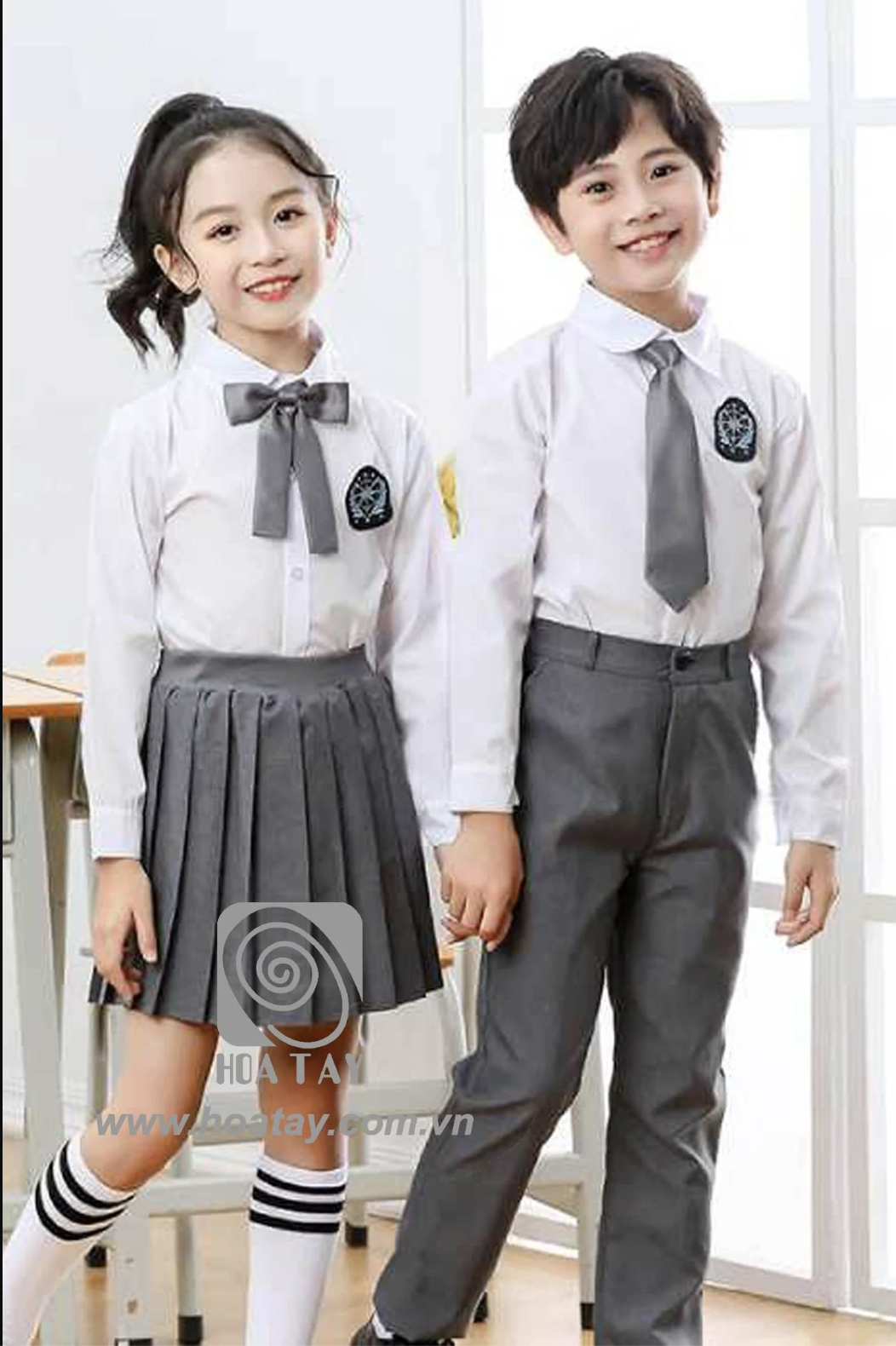 Hai học sinh (một nam, một nữ) đang đứng; mặc áo sơ mi trắng dài tay, nữ mặc chân váy xếp ly (xếp nếp) xám, nam mặc quần tây xám, thắt cà vạt/nơ xám, huy hiệu thêu trên ngực; bối cảnh lớp học, ánh sán