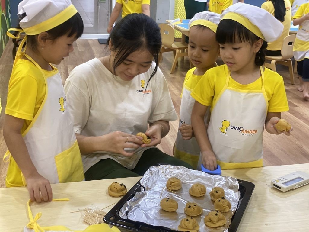 Ba trẻ em và một giáo viên đang làm bánh; họ đội mũ đầu bếp (mũ vải trắng) và đeo tạp dề (yếm bảo vệ) màu vàng trắng, thực hiện thao tác nặn bột trên khay nướng nhôm trong lớp học sáng sủa.