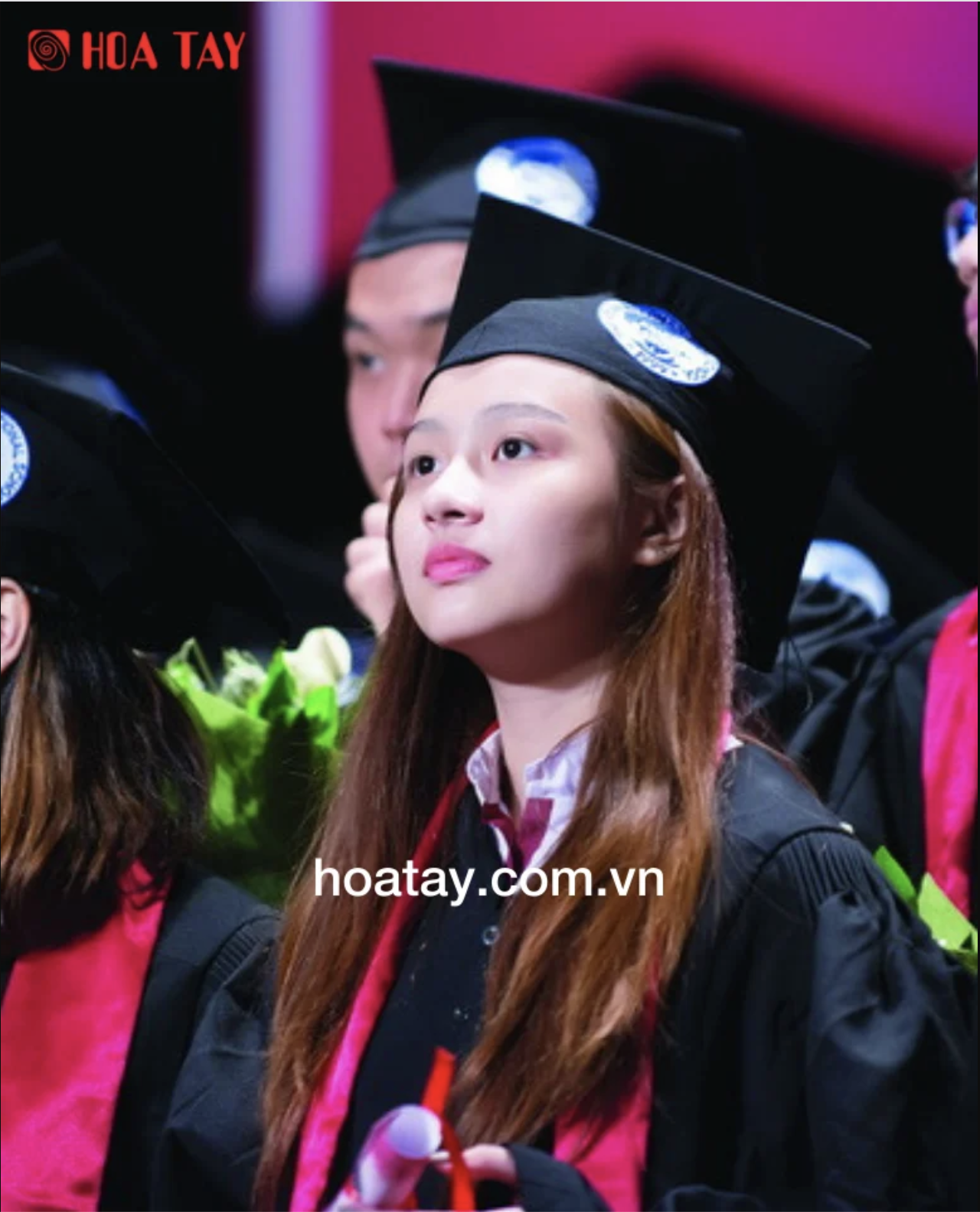 Một nữ sinh viên đang ngồi, đội mũ cử nhân (mũ tốt nghiệp vuông) màu đen, khoác áo lễ phục (áo cử nhân) màu đen với dải khăn choàng cổ màu đỏ, bối cảnh buổi lễ tốt nghiệp trong nhà với ánh sáng sân kh