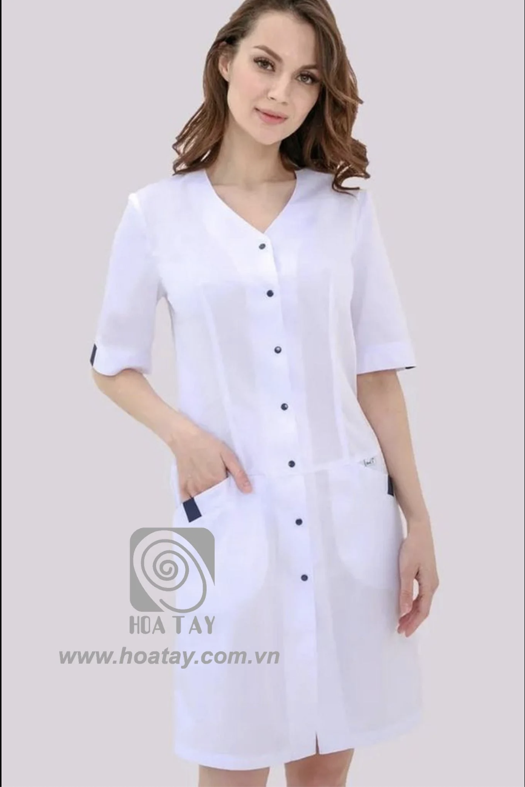 Một nữ nhân viên y tế đang đứng, mặc áo blouse trắng (áo khoác bác sĩ) cổ chữ V, cài khuy dọc (nút bấm), tay lỡ, có hai túi hông phối viền xanh đen, chất liệu vải phẳng mịn, phông nền studio sáng.