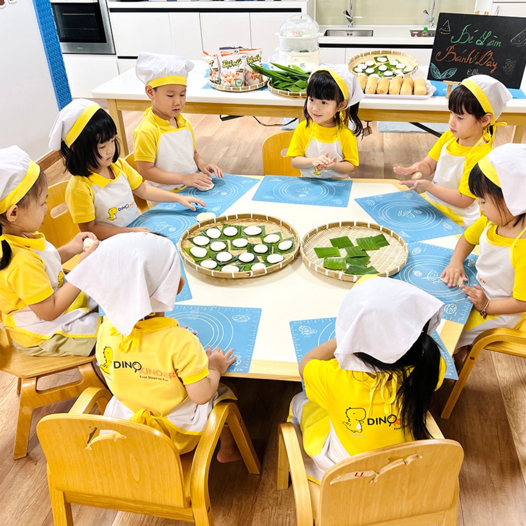 Tám trẻ mầm non đang làm bánh; đội mũ đầu bếp (mũ vải trắng) và tạp dề (yếm che ngực) màu vàng-trắng, ngồi quanh bàn tròn với thảm nhào bột silicon xanh, trong lớp học sáng sủa.