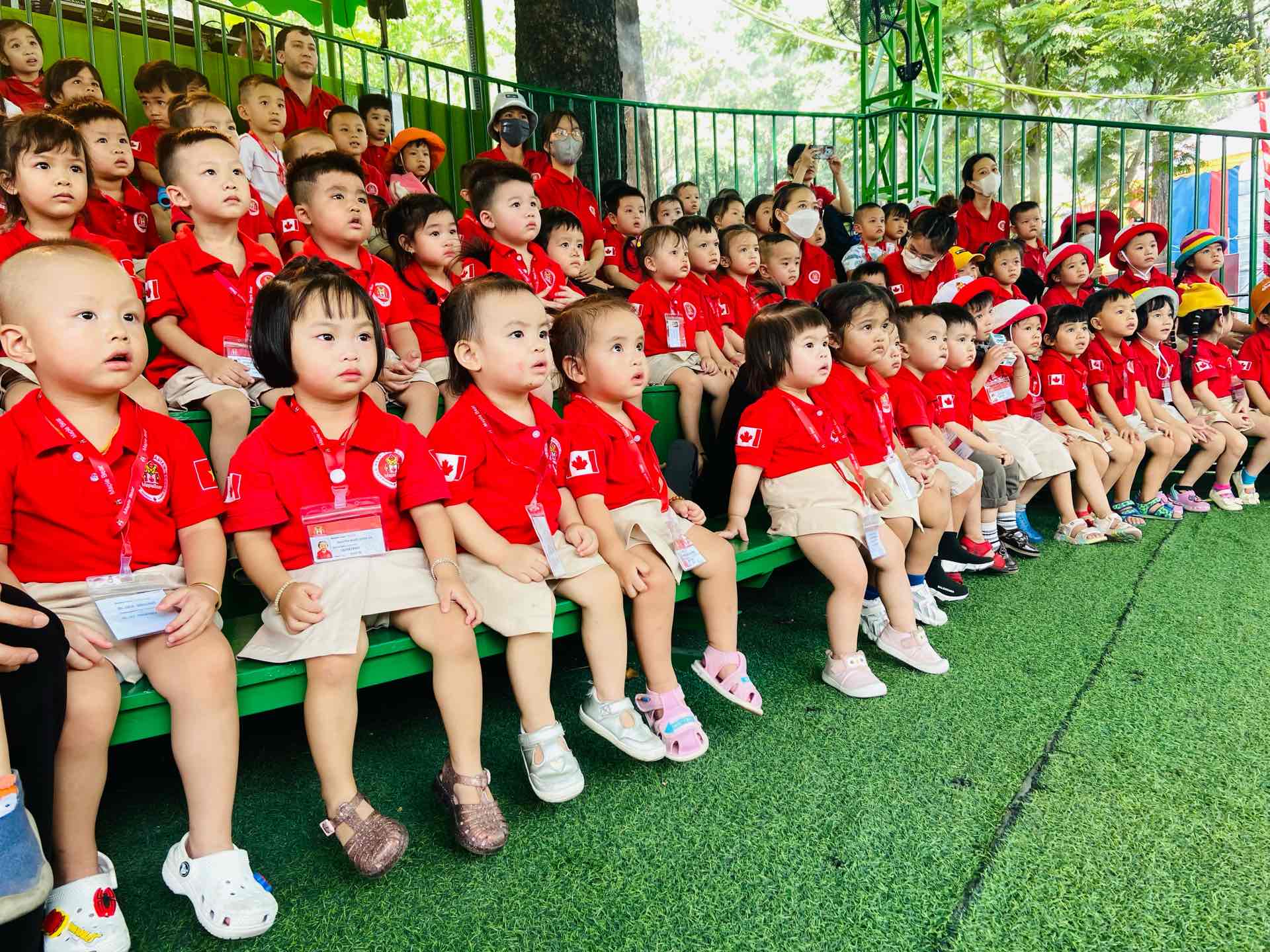 Một nhóm trẻ mầm non (khoảng 40 bé) đang ngồi trên khán đài, mặc áo polo đỏ (áo cổ bẻ) đồng phục, quần hoặc váy kaki màu be, đeo thẻ tên, dưới ánh sáng tự nhiên ngoài trời tại sân cỏ nhân tạo.