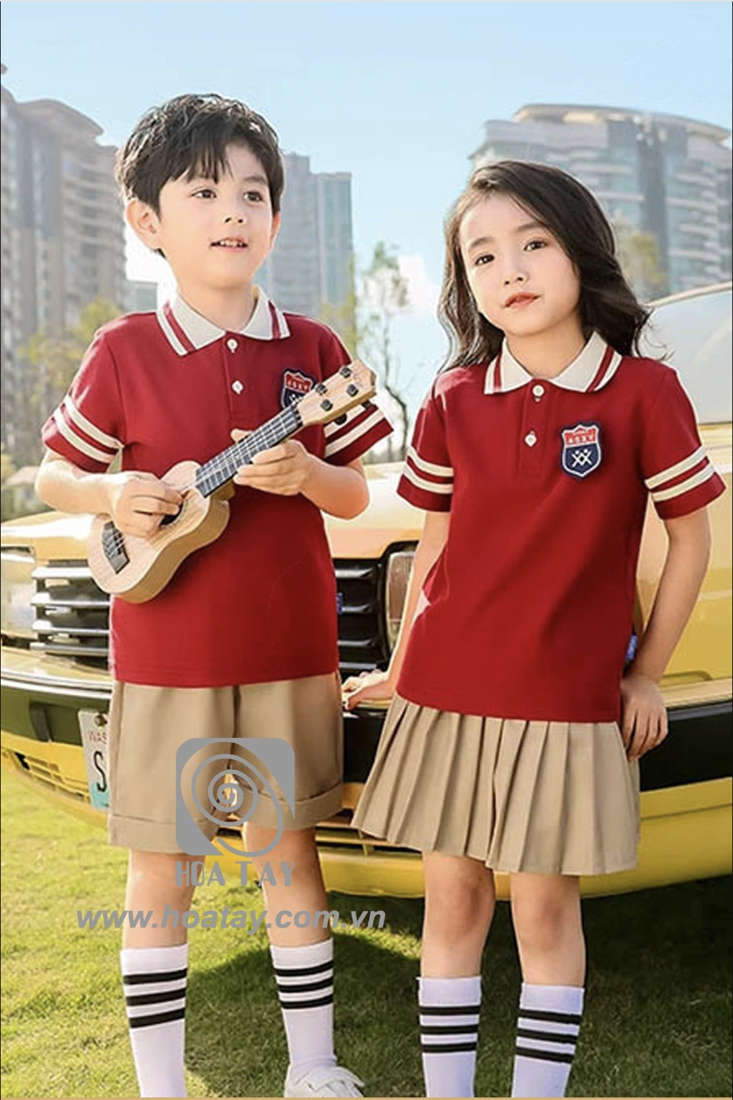 Hai trẻ em (một nam, một nữ) đang đứng tạo dáng, mặc áo polo đỏ cổ bẻ (cổ áo) phối viền trắng, quần short/váy xếp ly màu be, huy hiệu thêu trên ngực, tất cao cổ kẻ sọc, bối cảnh ngoài trời nắng.