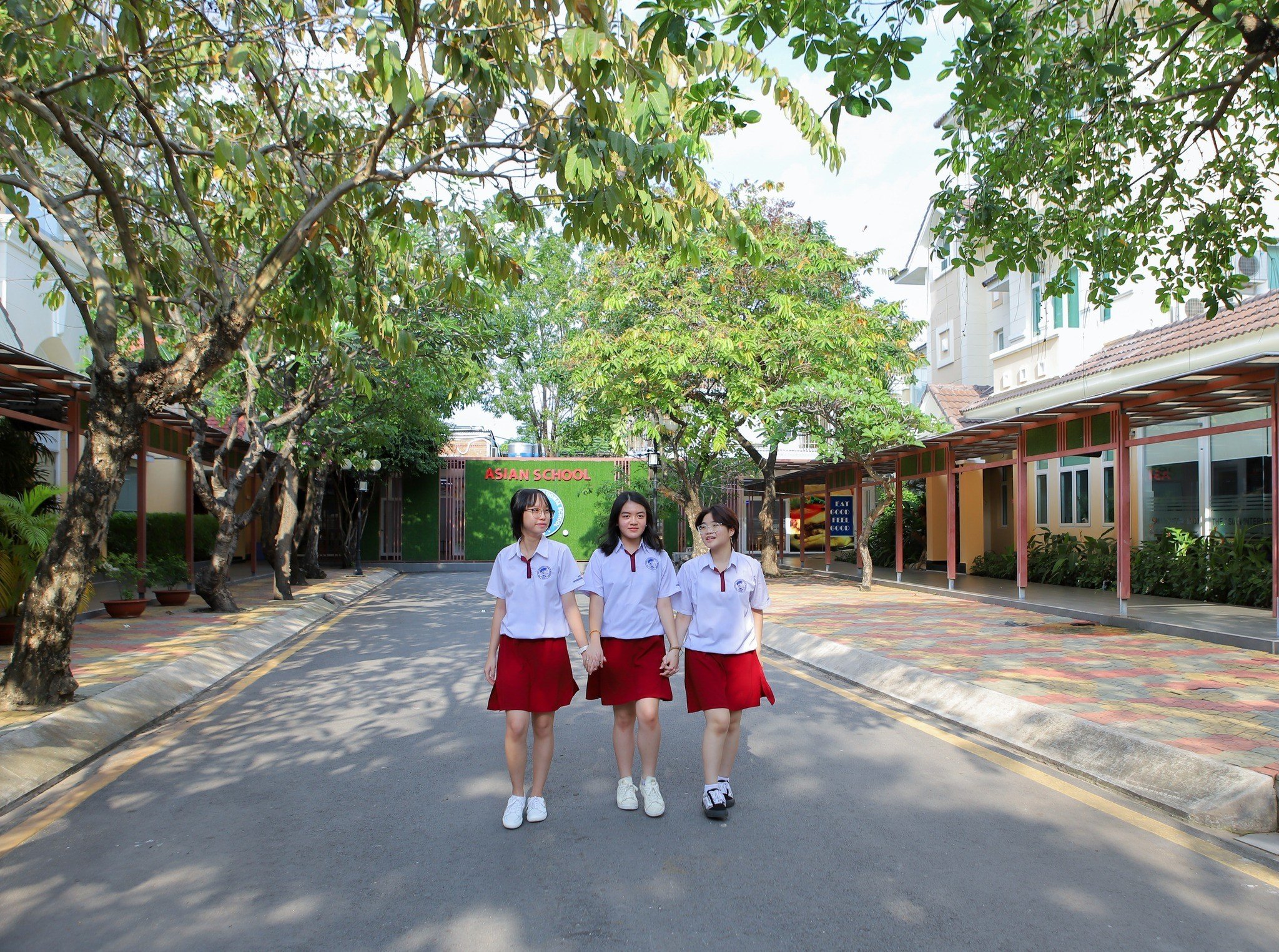 Ba nữ sinh đang đi bộ nắm tay nhau; mặc áo sơ mi trắng cổ bẻ (polo shirt) và chân váy đỏ (skirt) chất liệu vải thô, đi giày thể thao trắng; bối cảnh sân trường rợp bóng cây xanh, ánh sáng ban ngày tự