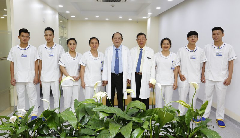 Nhóm 9 nhân viên y tế đứng trong hành lang bệnh viện. Họ mặc đồng phục trắng với viền cổ xanh, hai người đàn ông lớn tuổi mặc áo blouse trắng dài. Phía trước là chậu hoa diên vĩ trắng với lá xanh đậm.