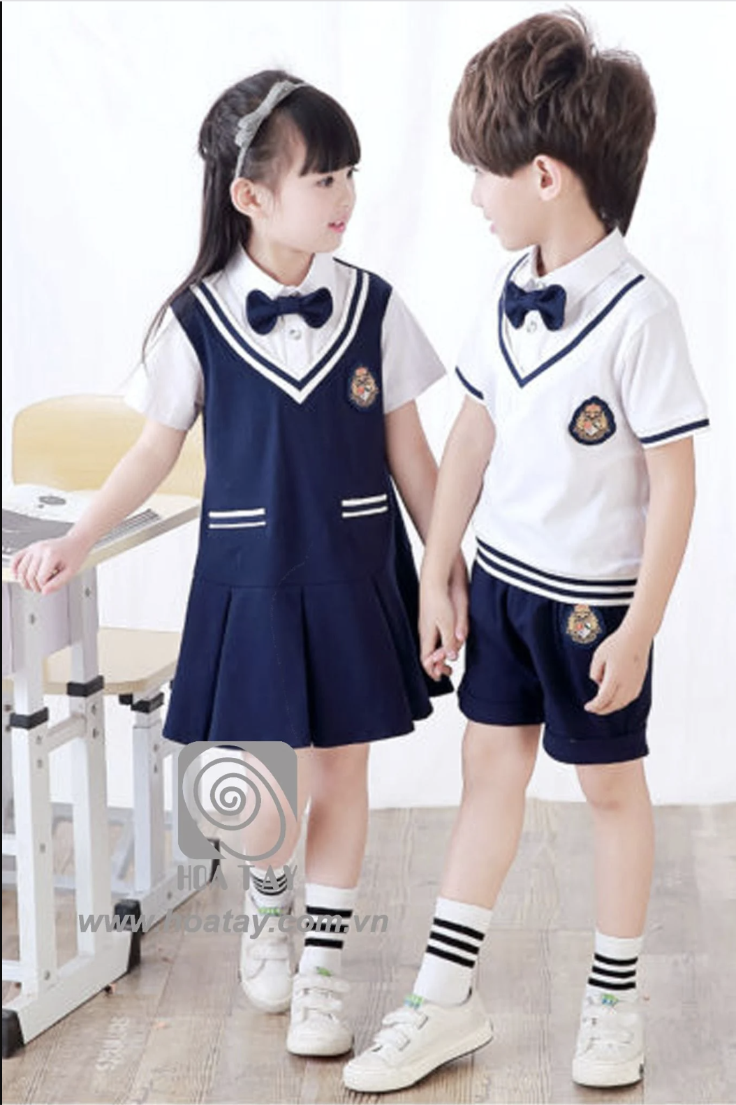 Hai học sinh (một nam, một nữ) đang đứng nắm tay nhau, mặc đồng phục cổ cồn (cổ bẻ) phối nơ, váy xếp ly và quần short xanh navy, huy hiệu thêu trên ngực, nền trắng sáng.