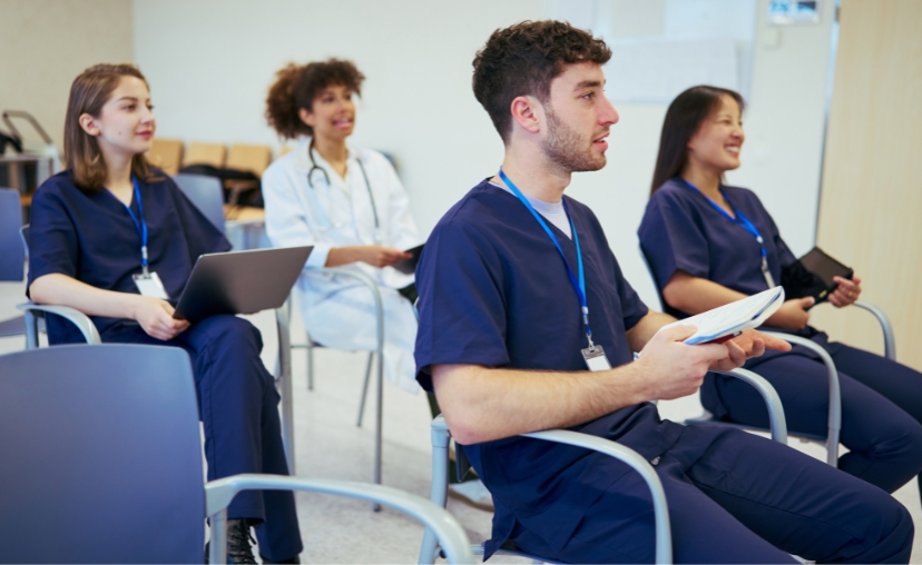Bốn nhân viên y tế ngồi trên ghế văn phòng, ba người mặc bộ đồng phục (scrubs) màu xanh navy, một người mặc áo blouse trắng với ống nghe. Họ cầm laptop và máy tính bảng, đeo thẻ nhân viên, trong phòng