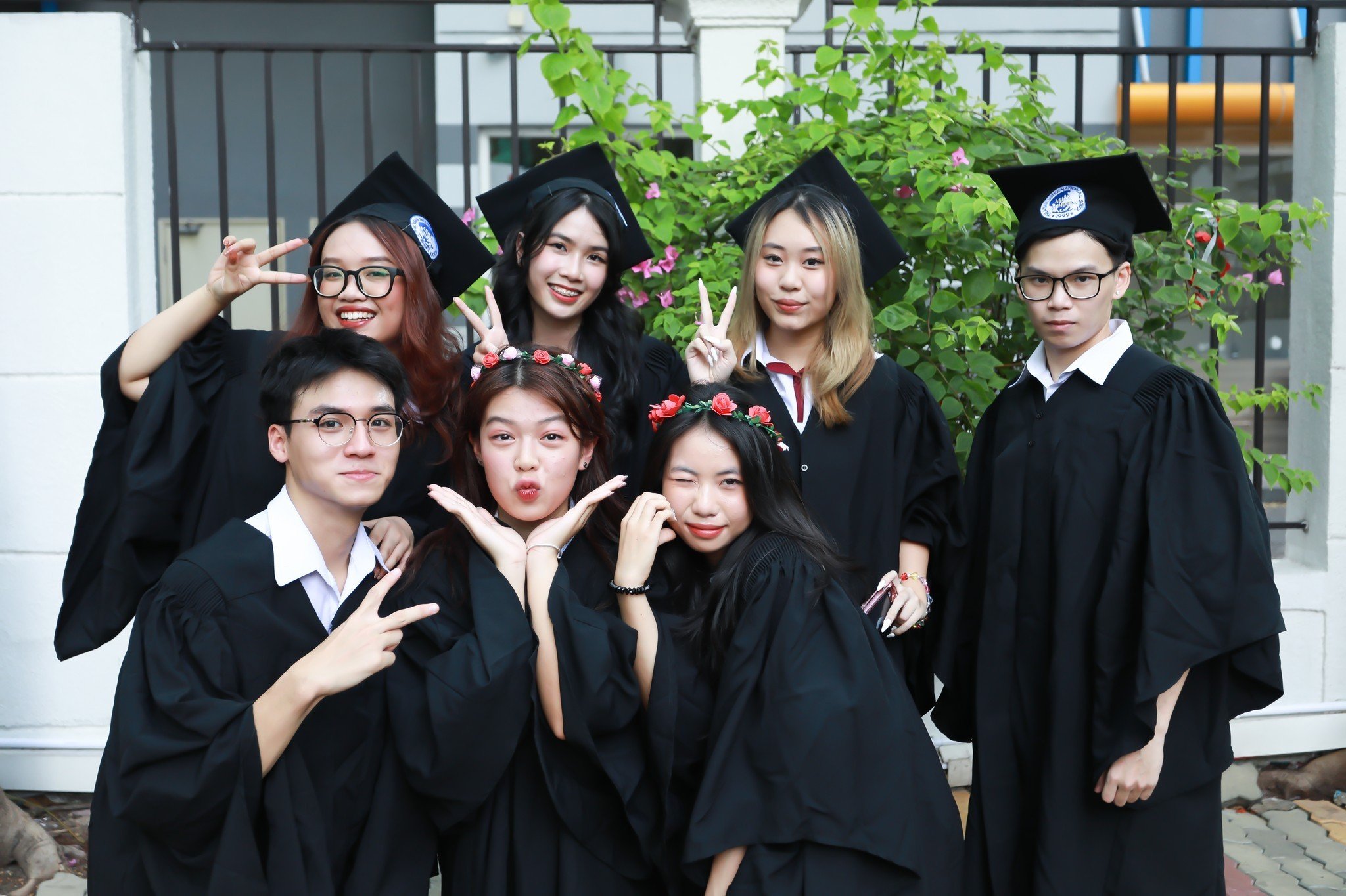 Nhóm 7 sinh viên đang tạo dáng chụp ảnh tốt nghiệp, mặc áo cử nhân (áo choàng đen) và mũ mortarboard (mũ vuông), đeo kính, vòng hoa đội đầu, bối cảnh ngoài trời với hàng rào sắt và cây xanh, ánh sáng
