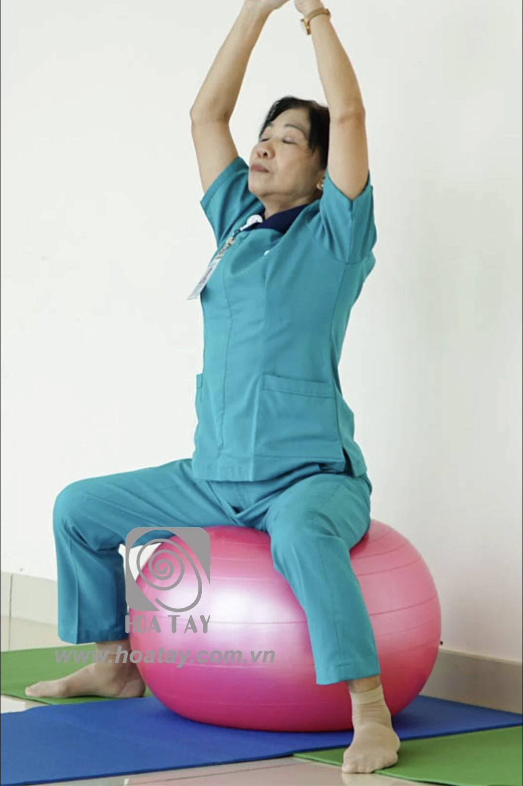 Một phụ nữ trung niên đang tập yoga trên bóng hơi (bóng tập gym) màu hồng; mặc bộ đồ y tá (scrubs) màu xanh cổ vịt, áo cổ chữ V có túi trước, quần ống đứng; không gian trong nhà, ánh sáng tự nhiên.