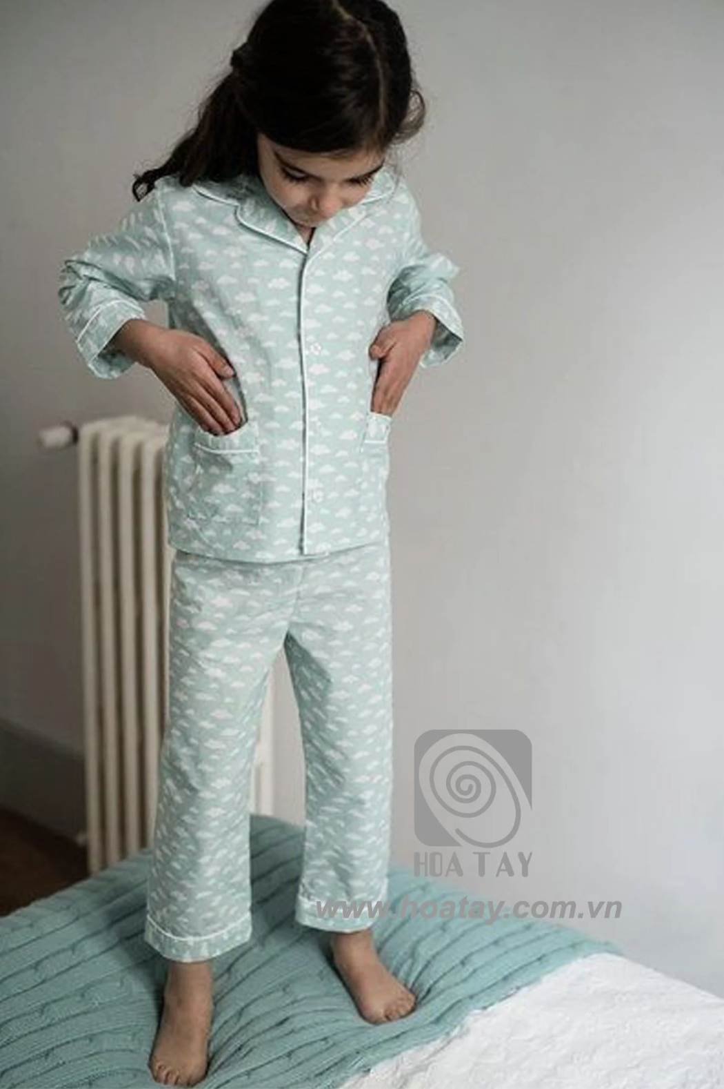 Một bé gái đang đứng trên giường, mặc bộ đồ ngủ (pajamas) màu xanh bạc hà họa tiết đám mây trắng, cổ bẻ (revere collar), cài cúc phía trước, có túi đắp, chất liệu cotton mềm, ánh sáng tự nhiên trong p