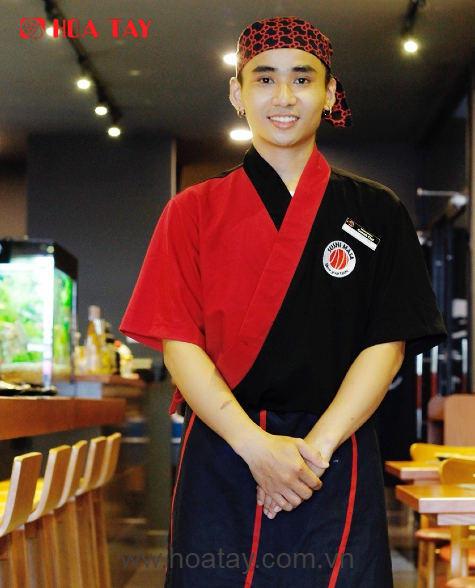 Một nam đầu bếp (đầu bếp nam) đang đứng mỉm cười; mặc áo khoác kimono (áo vạt chéo) phối hai màu đỏ đen, khăn bandana họa tiết, tạp dề đen viền đỏ; bối cảnh nhà hàng sushi, ánh sáng đèn trần trong nhà