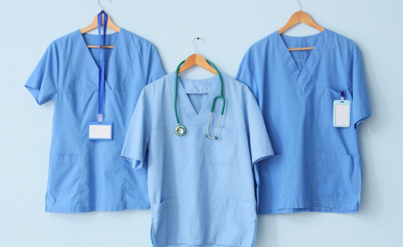 Ba bộ đồng phục y tá (scrubs) màu xanh dương, cổ chữ V, tay ngắn, treo trên móc gỗ. Hai bộ có dây đeo thẻ nhân viên, một bộ có ống nghe y tế màu xanh lá treo trước ngực. Nền tường màu xanh nhạt, ánh s