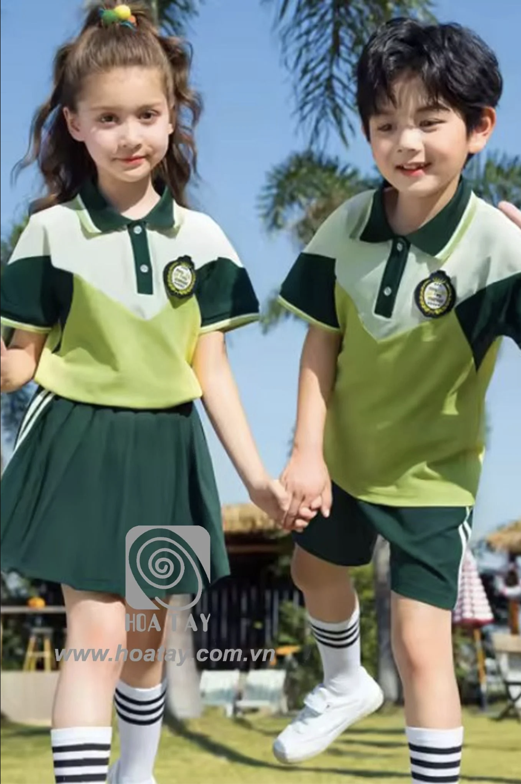 Hai trẻ em (một bé trai, một bé gái) đang nắm tay nhau; mặc đồng phục học sinh phối màu xanh lá và kem; áo polo cổ bẻ (cổ gập), chân váy xếp ly và quần short, huy hiệu thêu trên ngực; bối cảnh ngoài t