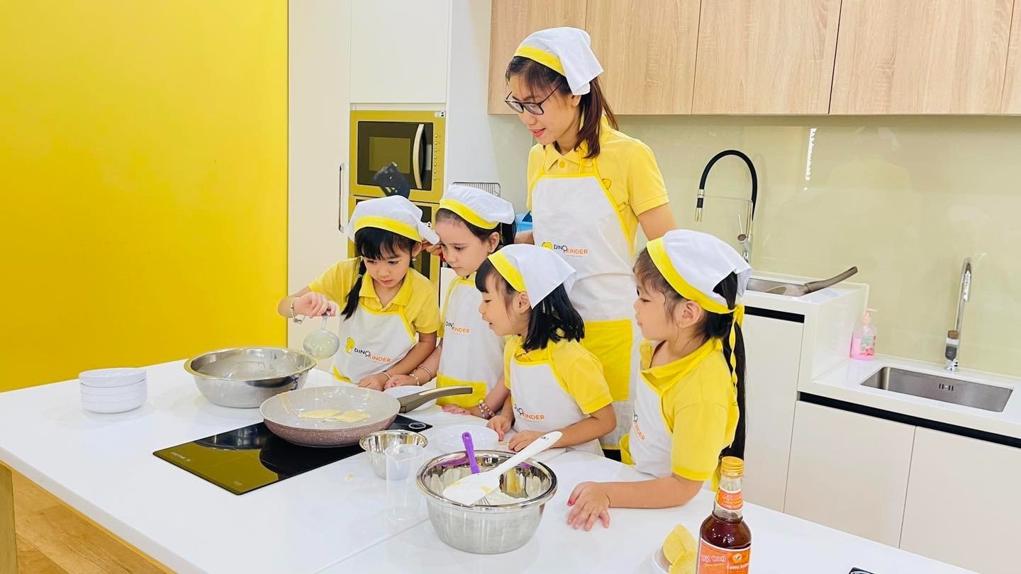 Một người lớn và bốn trẻ em đang nấu ăn; mặc áo thun vàng, tạp dề trắng (tấm vải che trước ngực) và mũ đầu bếp vải; bối cảnh bếp hiện đại, ánh sáng tự nhiên.