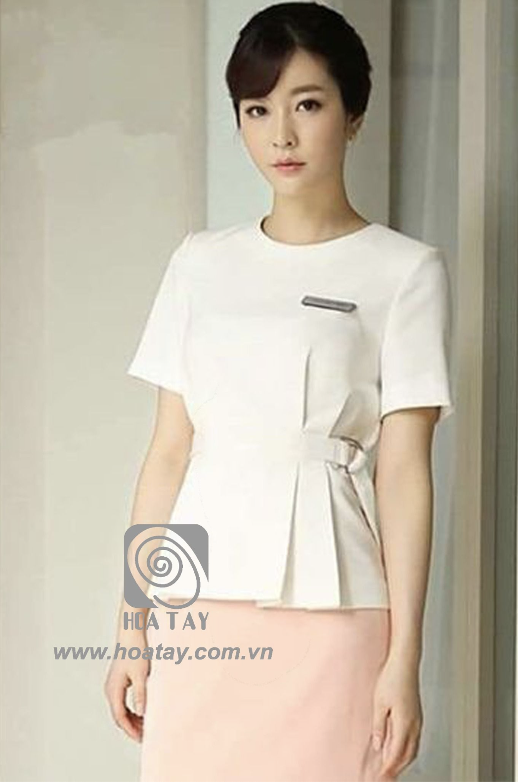 Một nữ nhân viên đang đứng, mặc áo đồng phục trắng cổ tròn (round neck), tay ngắn, chi tiết xếp ly (pleats) bên hông với đai thắt khóa cài (buckle belt), chất liệu vải đứng form, ánh sáng studio dịu n