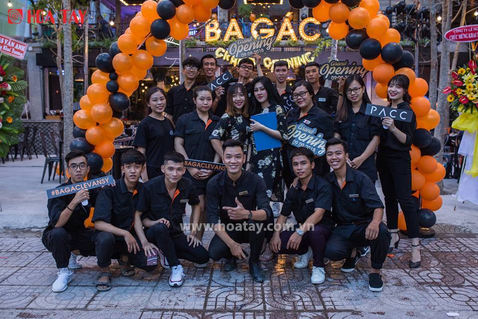 Nhóm 15 nhân viên (team) đang tạo dáng chụp ảnh; mặc đồng phục áo sơ mi đen (black shirt) cổ bẻ, chất vải cotton, phối viền cam; bối cảnh lễ khai trương (grand opening) với cổng bóng bay cam đen, ánh
