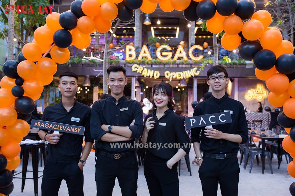 Bốn nhân viên (bốn người) đứng tạo dáng dưới cổng chào bong bóng cam đen, mặc áo sơ mi đen cổ tàu (cổ đứng) tay lỡ, quần tây đen, đeo bảng tên, bối cảnh nhà hàng khai trương với đèn trang trí, ánh sán