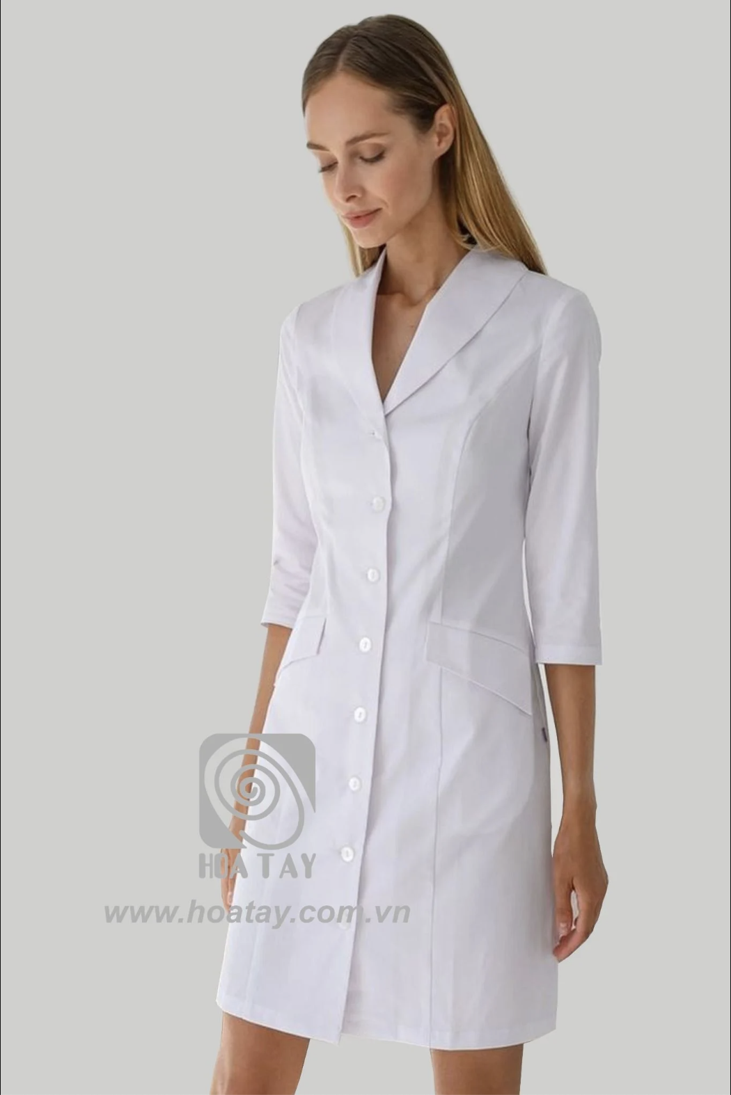 Một nữ nhân viên y tế đang đứng, mặc áo blouse trắng (áo khoác phòng thí nghiệm) tay lỡ, cổ bẻ (cổ vest), cài khuy đơn, có túi hông, chất liệu vải phẳng phiu, nền xám trung tính, ánh sáng studio.