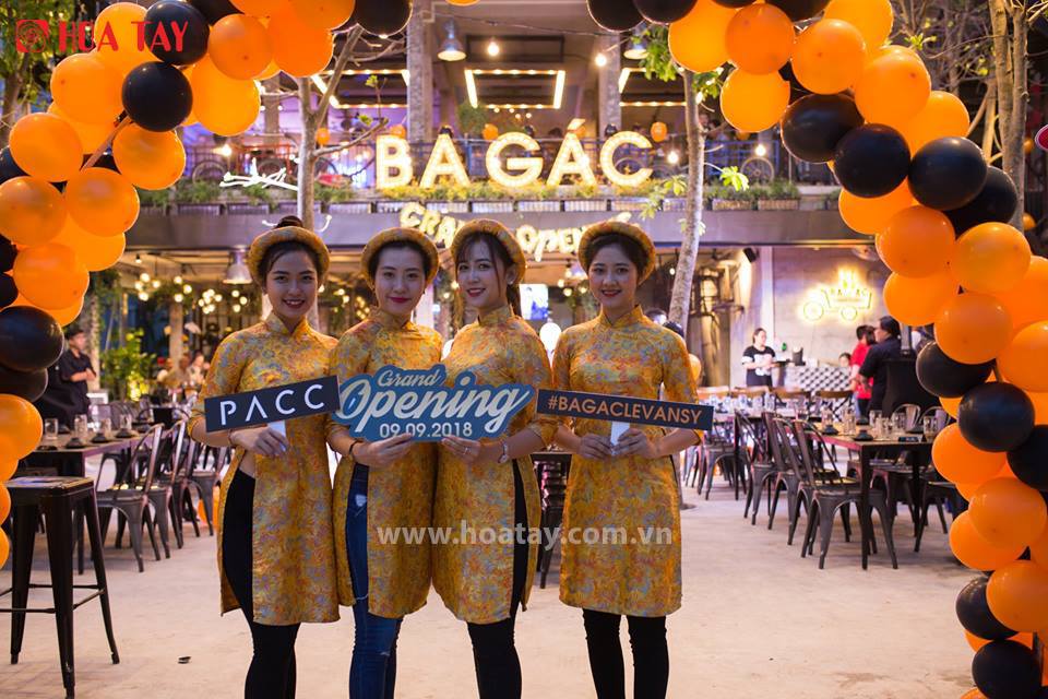 Bốn nữ nhân viên (bốn người) đang đứng mỉm cười, mặc áo dài (trang phục truyền thống) màu vàng họa tiết hoa, đội mấn (khăn vấn) đồng bộ, cầm bảng hiệu sự kiện, dưới cổng chào bóng bay cam đen tại khôn
