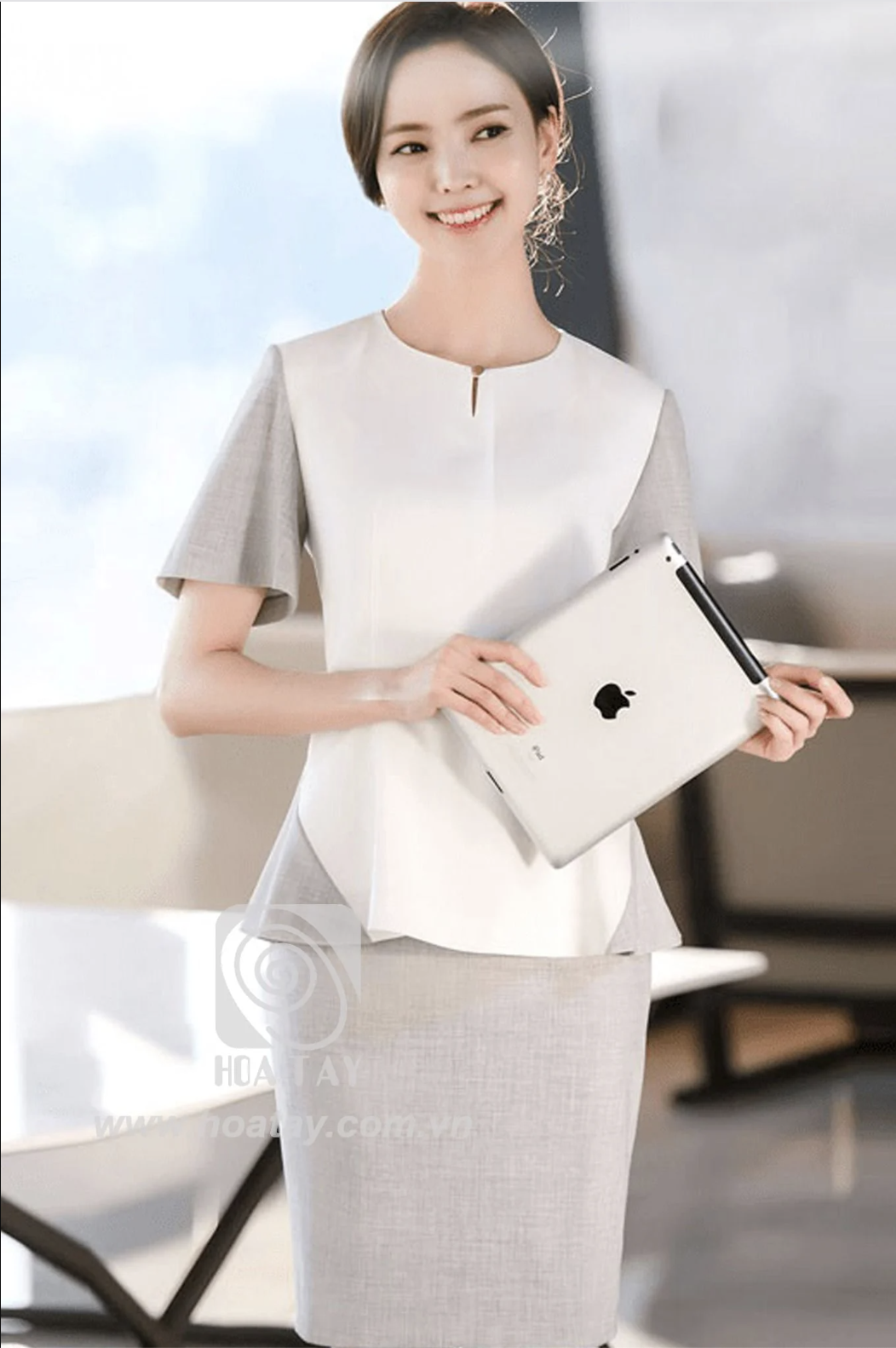 Một nữ nhân viên văn phòng đang đứng, mặc áo peplum phối màu trắng và xám, cổ tròn xẻ chữ V nhỏ, tay ngắn, chân váy bút chì cùng tông màu, bối cảnh văn phòng sáng sủa.