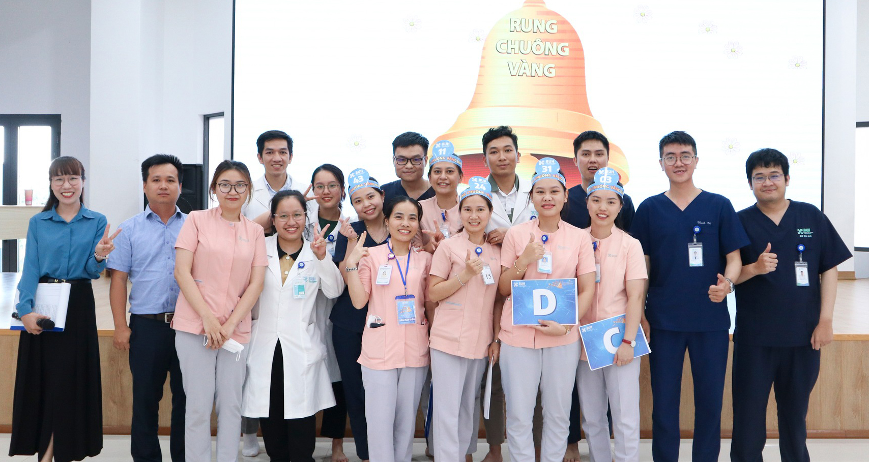 Nhóm 16 nhân viên y tế đang tạo dáng chụp ảnh, một số đội vương miện giấy (mũ giấy) ghi số, mặc áo blouse trắng hoặc đồng phục y tá màu hồng nhạt, đeo thẻ tên, đứng trước màn hình chiếu hình cái chuôn