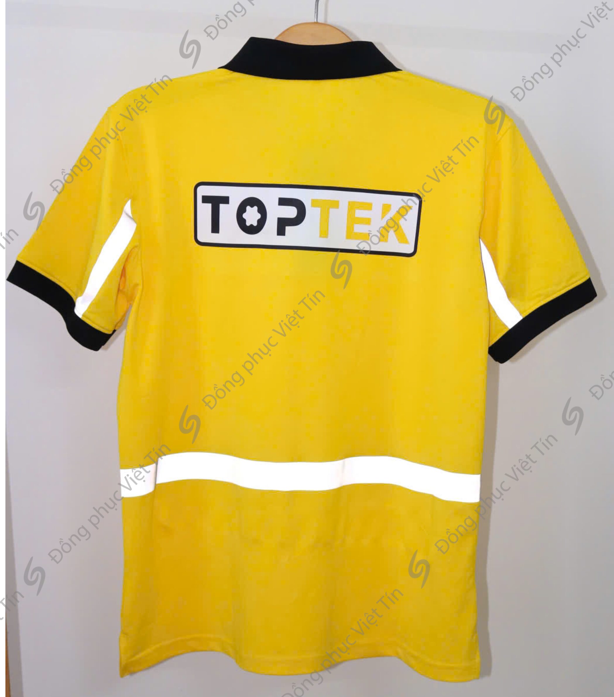 Áo thun đồng phục màu vàng tươi, cổ và tay áo màu đen, có dải phản quang ở tay và thân áo. Logo TOPTEK in nổi bật ở giữa lưng, treo trên móc áo trước nền trắng.