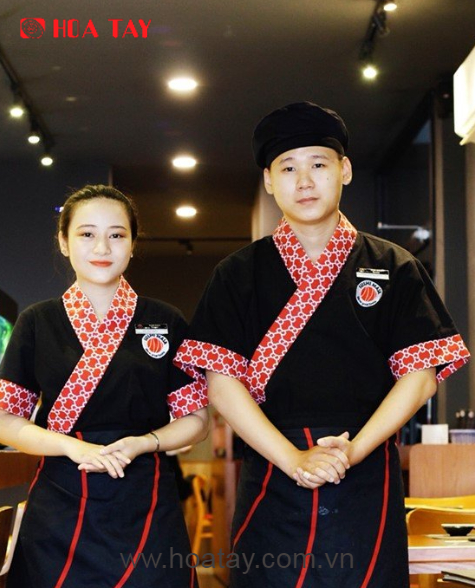 Hai nhân viên (một nam, một nữ) đang đứng thẳng, mặc áo đầu bếp kiểu Nhật (kimono) màu đen, cổ và tay áo phối họa tiết đỏ trắng, tạp dề đen viền đỏ, mũ nồi đen, bối cảnh nhà hàng với ánh sáng vàng ấm