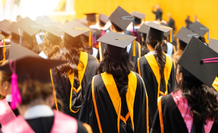 Nhóm sinh viên tốt nghiệp từ phía sau, đội mũ cử nhân (mortarboard) màu đen có tua rua, mặc áo choàng (gown) đen với dải viền vàng và hồng, đứng trong hội trường tốt nghiệp với ánh sáng ấm áp.