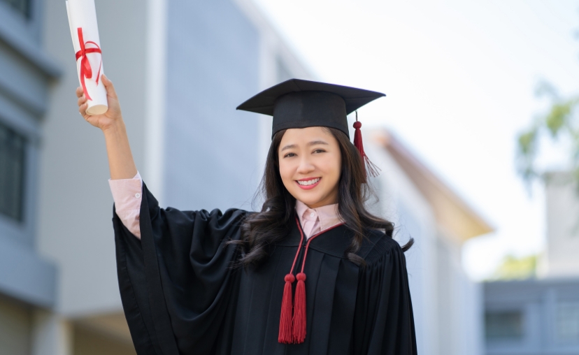 Một nữ sinh viên châu Á -> giơ cao bằng tốt nghiệp cuộn trắng có nơ đỏ -> mũ cử nhân đen có tua rua đỏ, áo toga đen, áo sơ mi hồng nhạt bên trong -> ngoài trời, ánh sáng ban ngày, tòa nhà mờ ở hậu cản