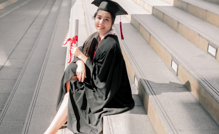 Một nữ sinh viên tốt nghiệp ngồi trên bậc thềm đá ngoài trời, mặc áo choàng (gown) đen dài và mũ tốt nghiệp (mortarboard) có tua rua đỏ, tay cầm cuộn bằng tốt nghiệp (diploma) buộc nơ đỏ, ánh sáng mặt