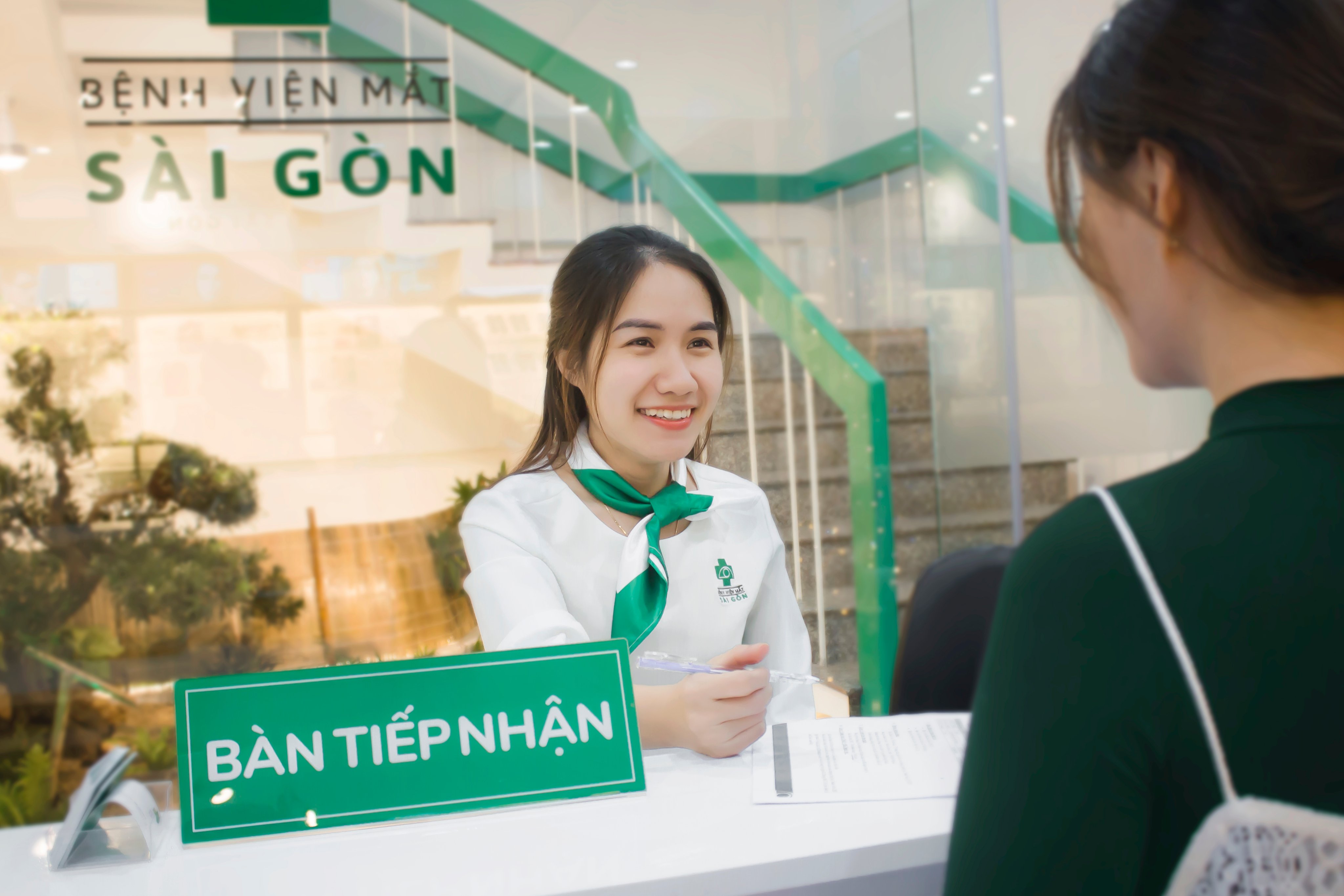 Nhân viên nữ mặc áo sơ mi trắng, khăn lụa xanh lá, ngồi sau quầy tiếp tân có biển 'BÀN TIẾP NHẬN', trao giấy tờ cho khách, nền là cầu thang kính xanh và logo bệnh viện mắt.