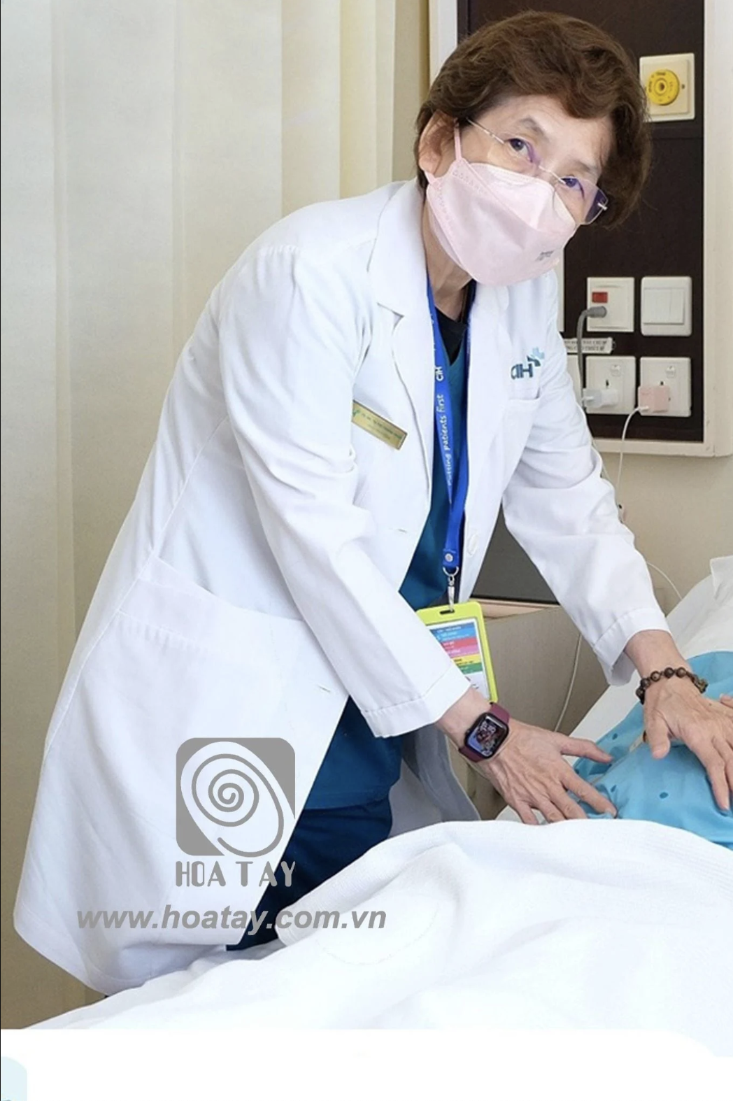 Một nữ bác sĩ đang thăm khám bệnh nhân; mặc áo blouse trắng (áo khoác y tế) cổ bẻ, tay dài, chất liệu vải cotton dày, có bảng tên và logo thêu ngực, đeo khẩu trang y tế màu hồng, bối cảnh phòng bệnh v