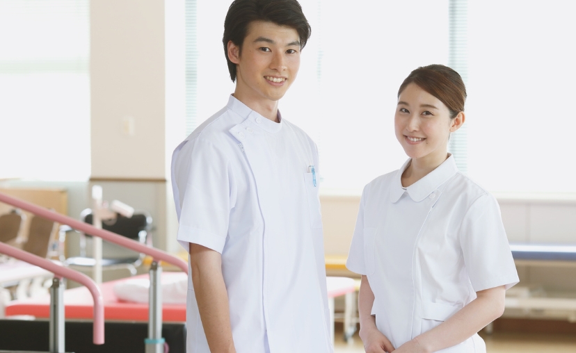 Hai nhân viên y tế (một nam, một nữ) đứng trong phòng bệnh viện. Họ mặc đồng phục trắng: áo nam cổ đứng, áo nữ cổ bẻ. Nền có giường bệnh và lan can hồng, ánh sáng tự nhiên.