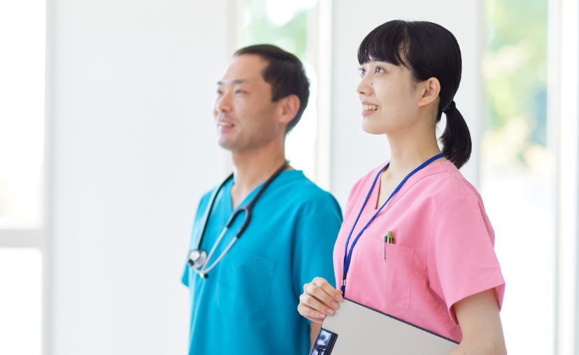 Hai bác sĩ (một nam, một nữ) đứng cạnh nhau trong hành lang bệnh viện. Người nam mặc áo blouse xanh dương, đeo ống nghe. Người nữ mặc áo blouse hồng, cầm bảng ghi chú. Ánh sáng tự nhiên, nền mờ.